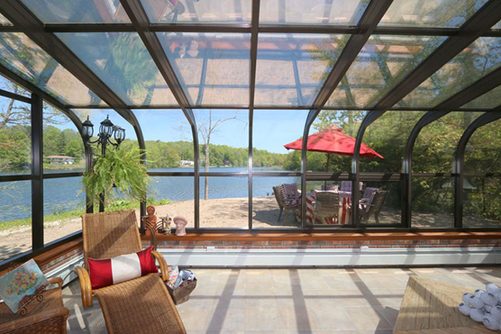 Interior of a glass room with wall and ceiling windows and dark brown framing, a lounge chair and lantern inside, and a patio with an outdoor table and chairs with an umbrella over it and a lake outside.